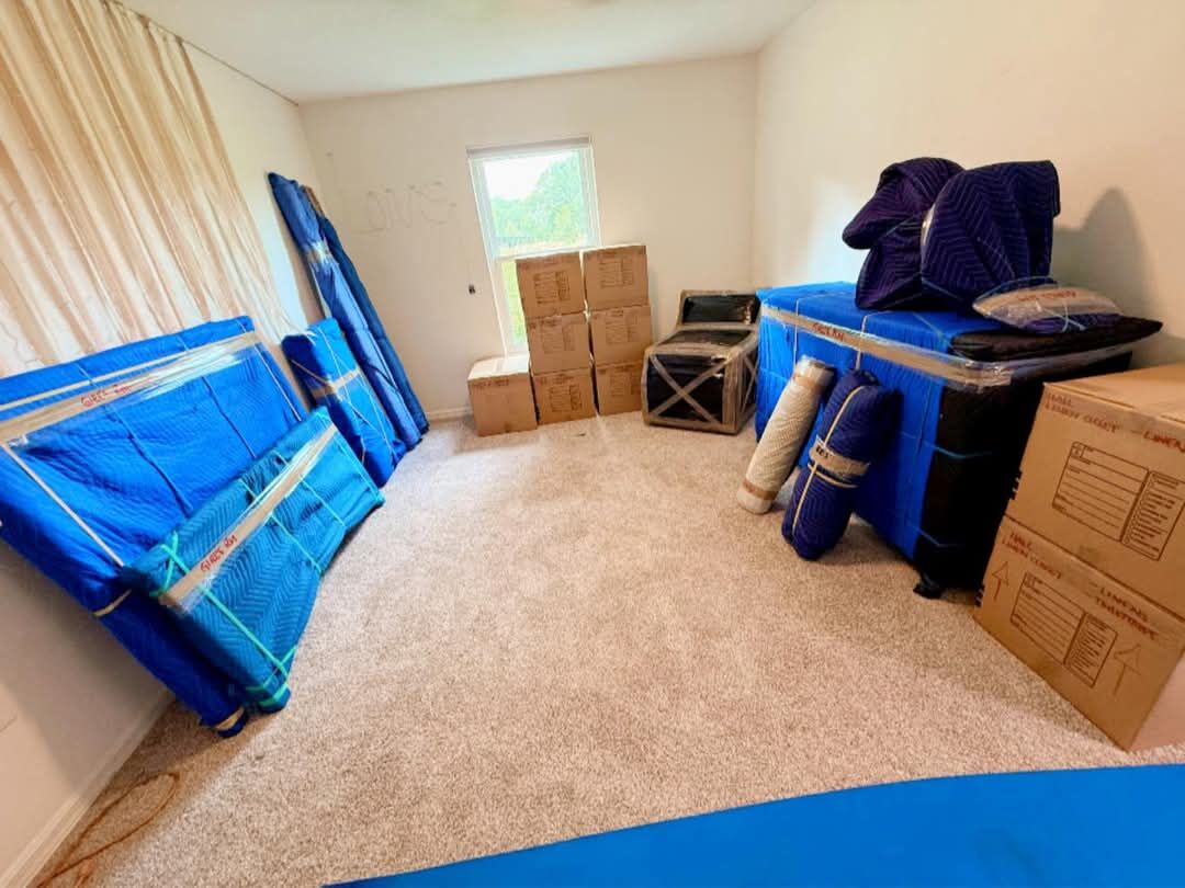 Bedroom with blue blanket-wrapped bed frame, rolled rugs, crated item and boxes staged for move day — One World Van Lines Wylie, TX DFW professional movers