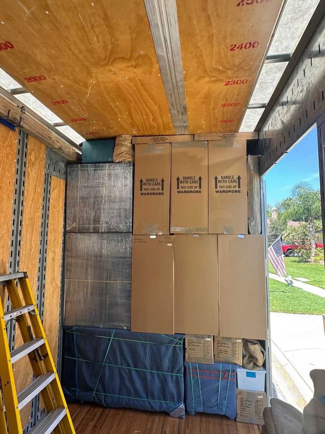Wardrobe Handle-With-Care boxes and plastic-wrapped flat panels loaded in moving truck at palm-tree suburban home — One World Van Lines Cedar Park, Austin TX movers
