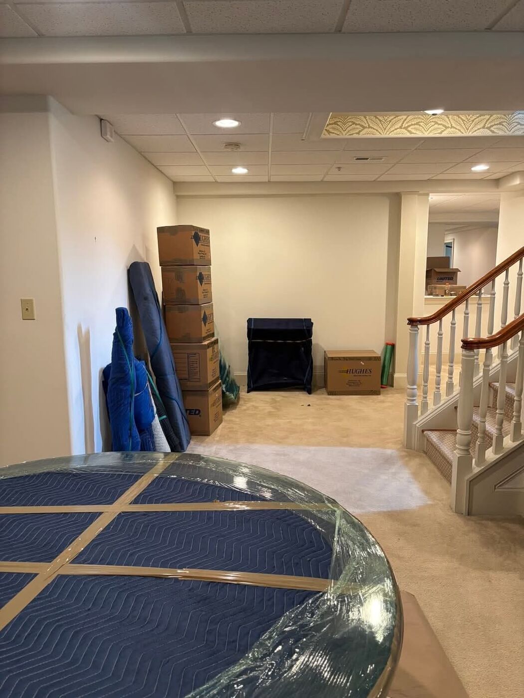 Wrapped round glass table top, stacked boxes and blanket-wrapped items in finished basement near staircase — One World Van Lines Rowlett, TX DFW movers